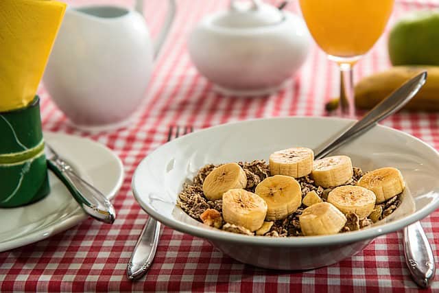Banana cereal breakfast bowl on a red checkered tablecloth with breakfast items and drinks in the background.